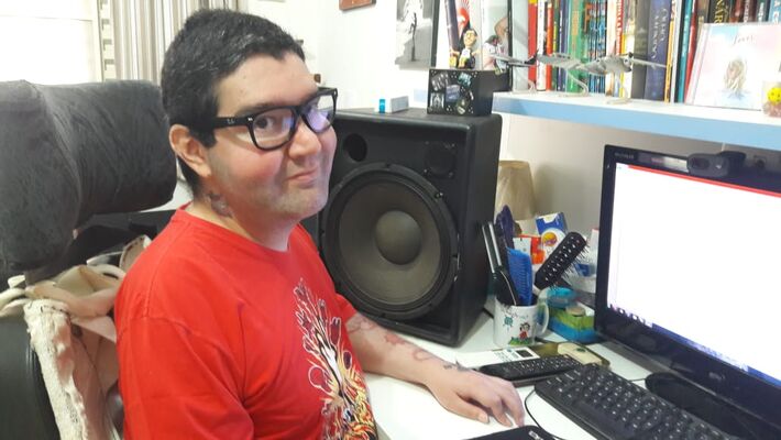 #PraCegoVer – Fotografia. Pedro sentado em frente a um computador. Ele tem cabelos pretos curtos, usa óculos e veste uma camiseta vermelha. Ao fundo, há uma estante com livros. Fim da Descrição.