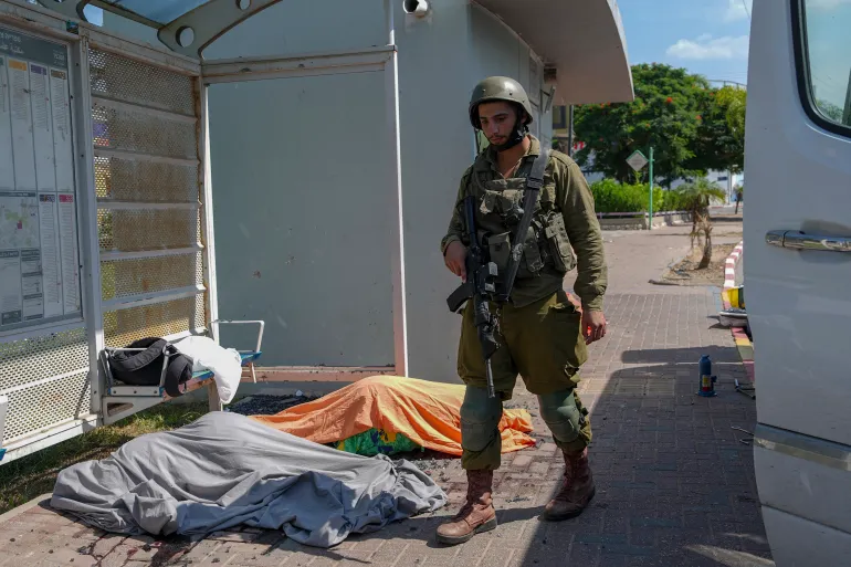 Um soldado israelense fica ao lado dos corpos de israelenses mortos por combatentes palestinos que entraram vindos da Faixa de Gaza. [Tsafrir Abayov/AP Photo]